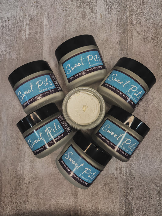 Six jars of Sweet Pits arranged in a circle on a textured gray surface, with one open jar in the center showing the creamy off-white oil.