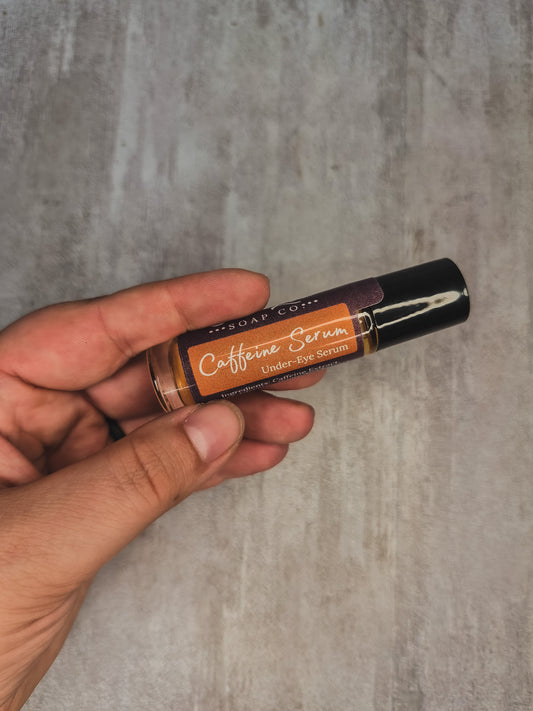 A hand holding a small cosmetic tube labeled 'Caffeine Serum' against a textured gray background.
