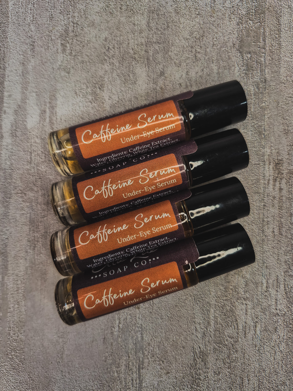 Four under-eye serum rollerball bottles from Soap Co. arranged diagonally on a textured gray background.