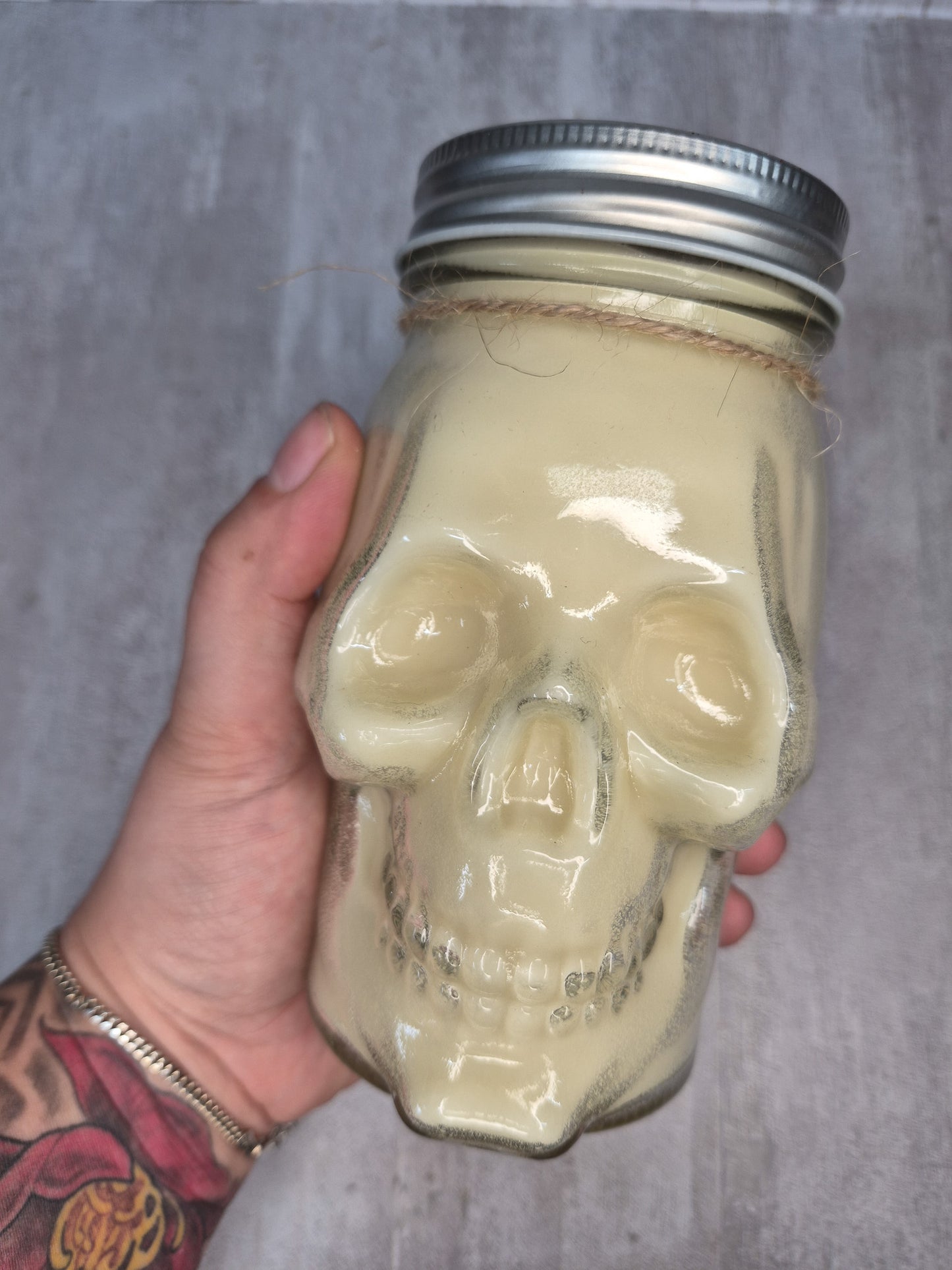 A hand holding a skull-shaped glass jar with a silver metal screw-top lid and a thin twine detail near the neck, against a gray textured background.