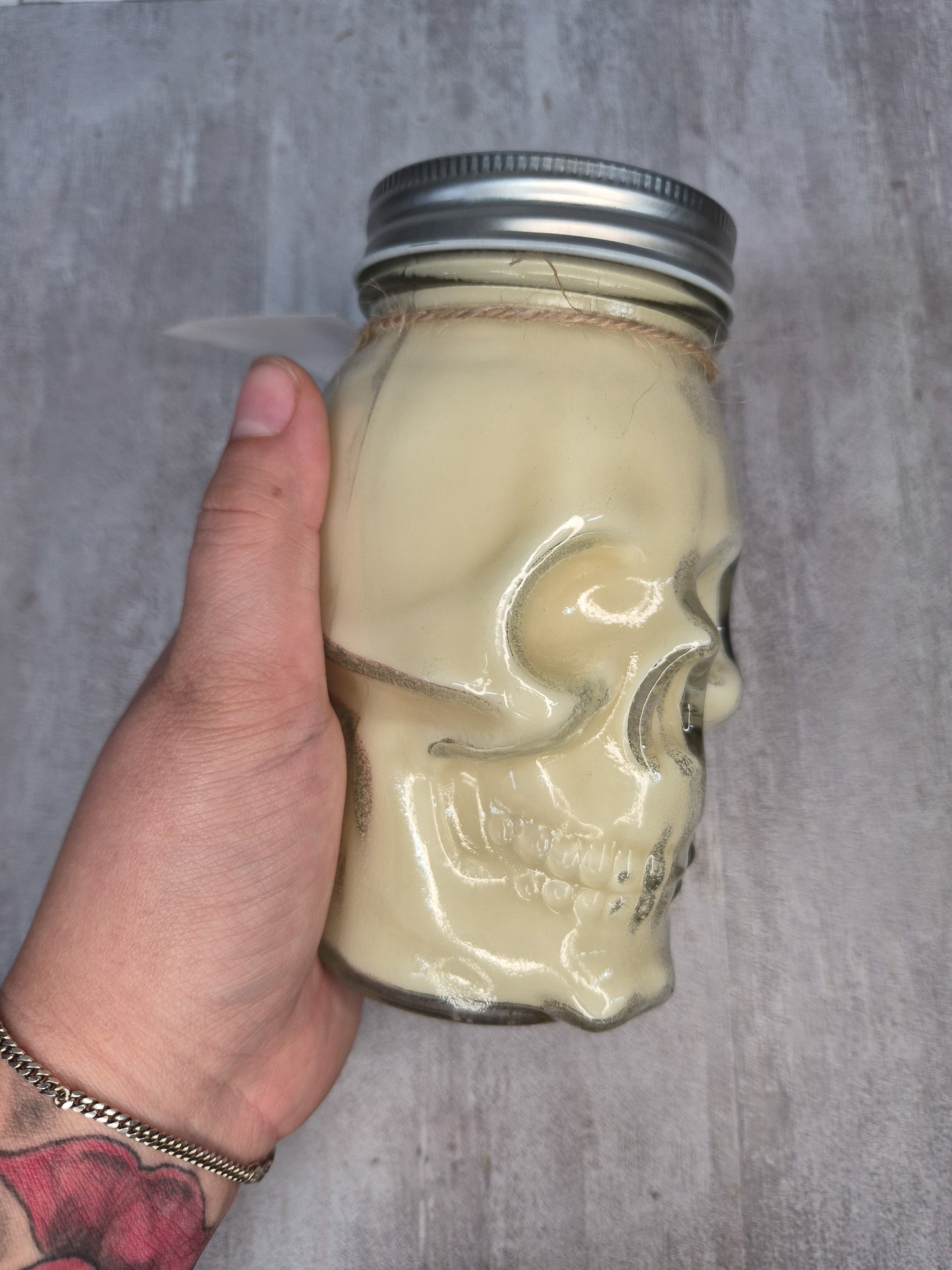 A hand holding a skull-shaped glass jar with a silver metal screw-top lid, set against a textured gray background.