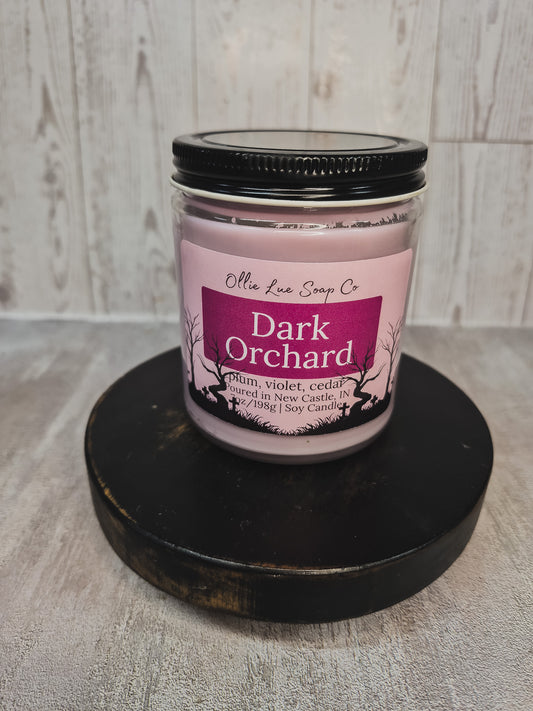 A jar candle labeled 'Dark Orchard' by Ollie Lue Soap Co, featuring a light pink wax and a black lid, displayed on a round black wooden riser against a light gray wood-plank background.