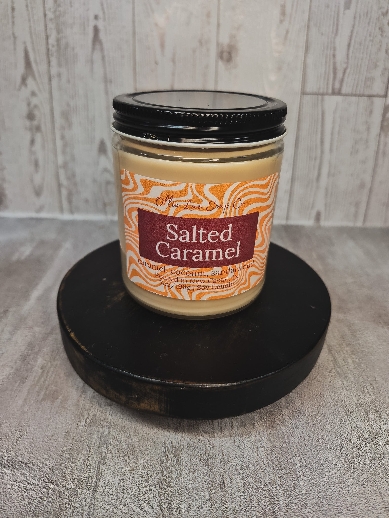 A jar candle labeled 'Salted Caramel' sits on a round black wooden platform against a light gray wood-plank background. The candle wax is a warm caramel beige, with an orange and white wavy patterned label and a black lid.