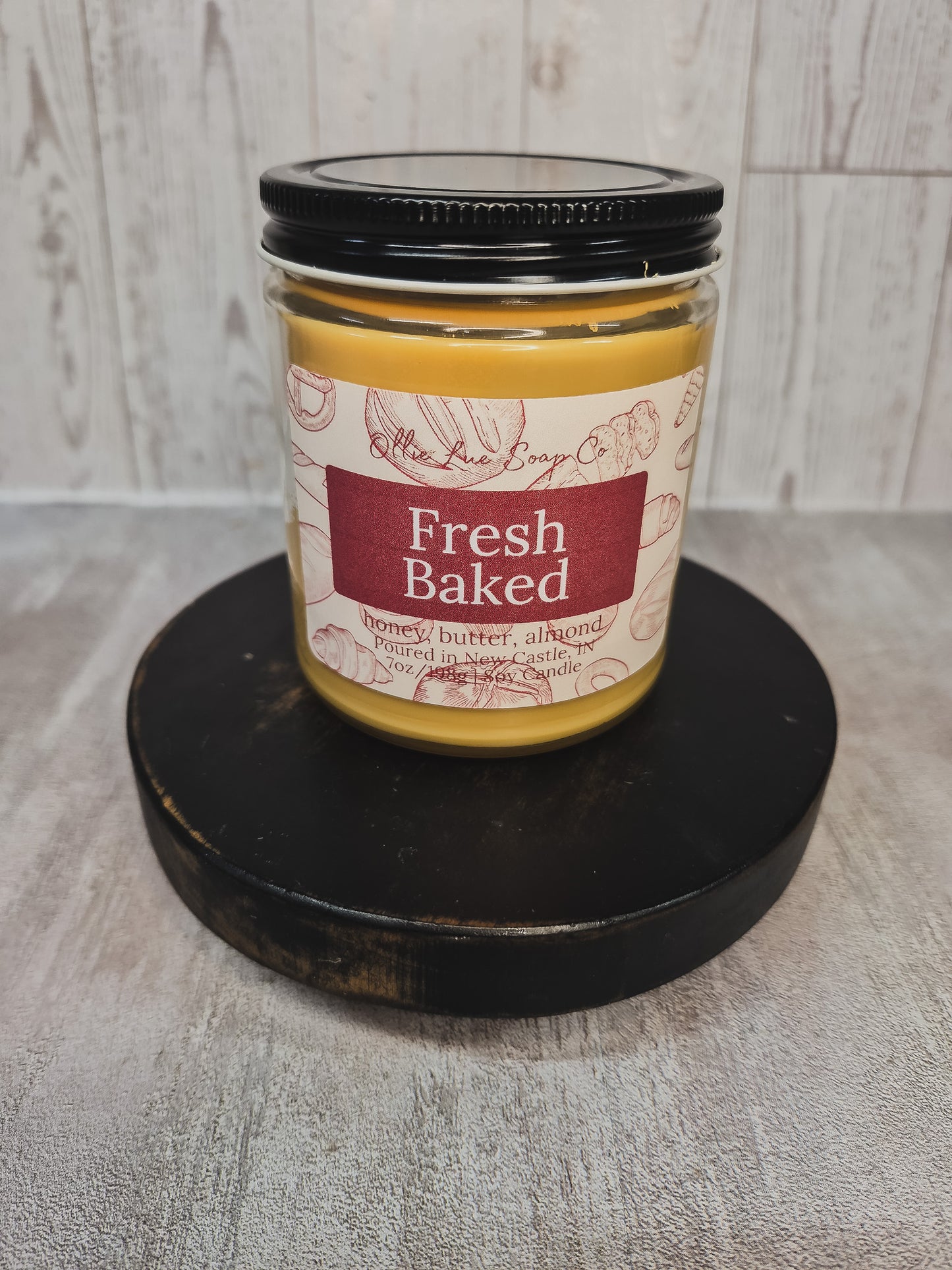 A jar candle labeled 'Fresh Baked' by Ollie Lue Soap Co., filled with yellow wax and topped with a black lid, displayed on a round black wooden riser against a light gray wood-plank background.