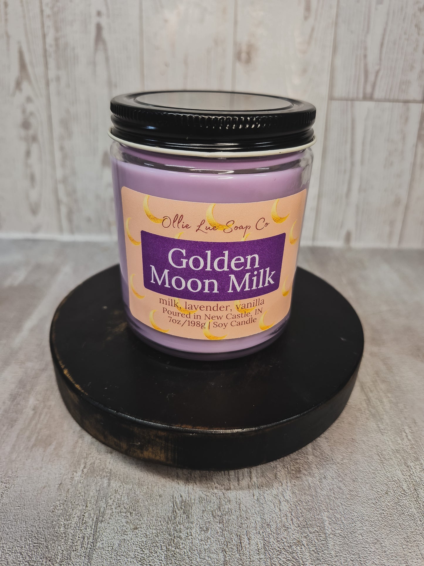 A lavender soy candle in a clear glass jar with a black lid, displayed on a round black wooden riser. The label reads 'Olie Lue Soap Co.