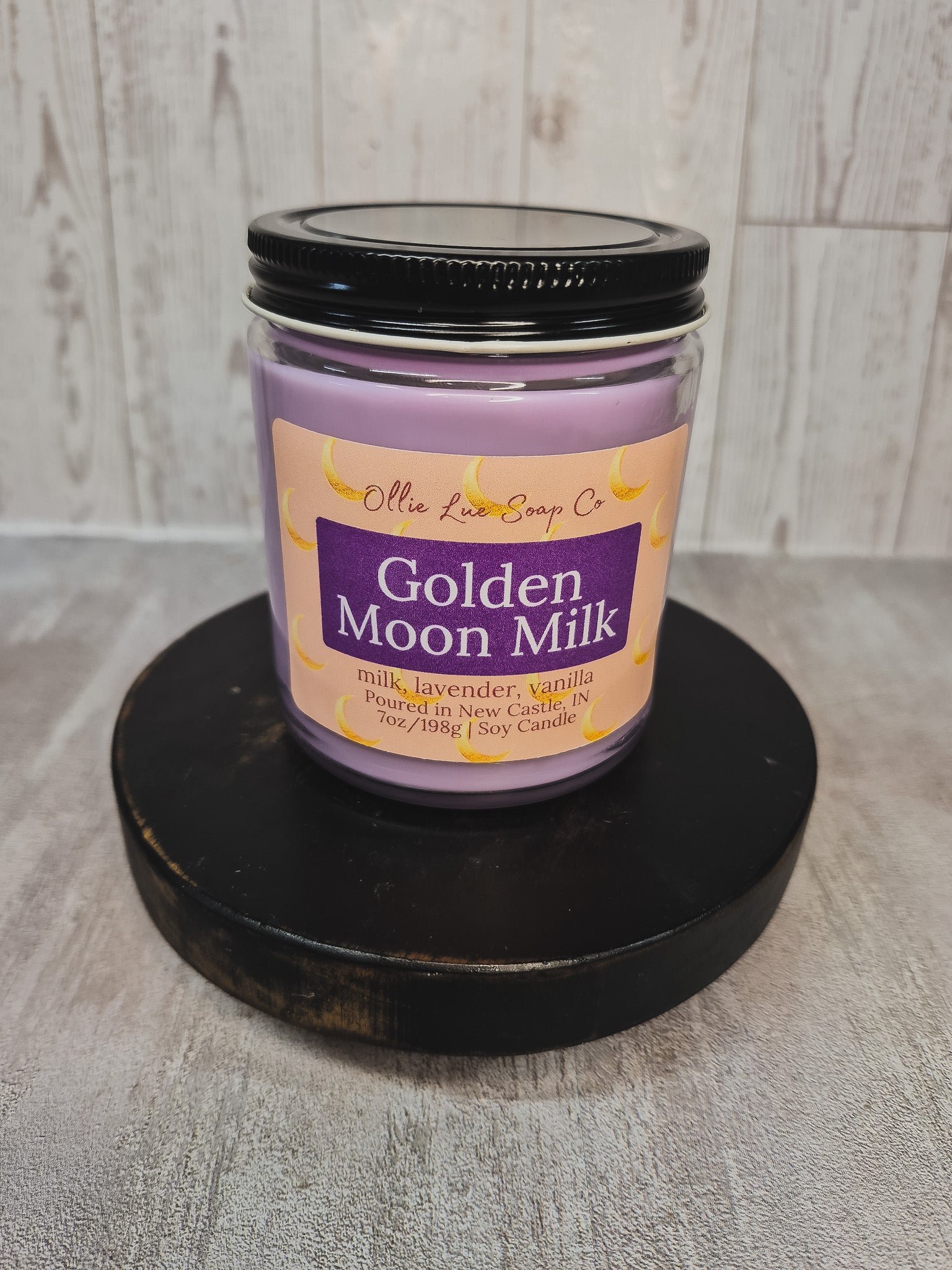 A lavender soy candle in a clear glass jar with a black lid, displayed on a round black wooden riser. The label reads 'Olie Lue Soap Co.