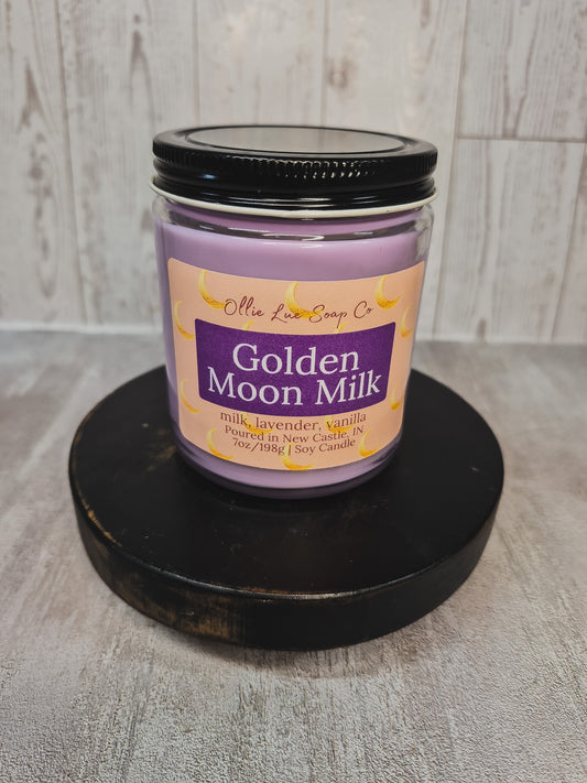 A lavender soy candle in a clear glass jar with a black lid, displayed on a round black wooden riser. The label reads 'Olie Lue Soap Co.