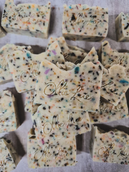 Multiple rectangular bars of handmade soap with colorful flecks and the brand name 'Ollie Lue' embossed on the front, arranged on a light surface.
