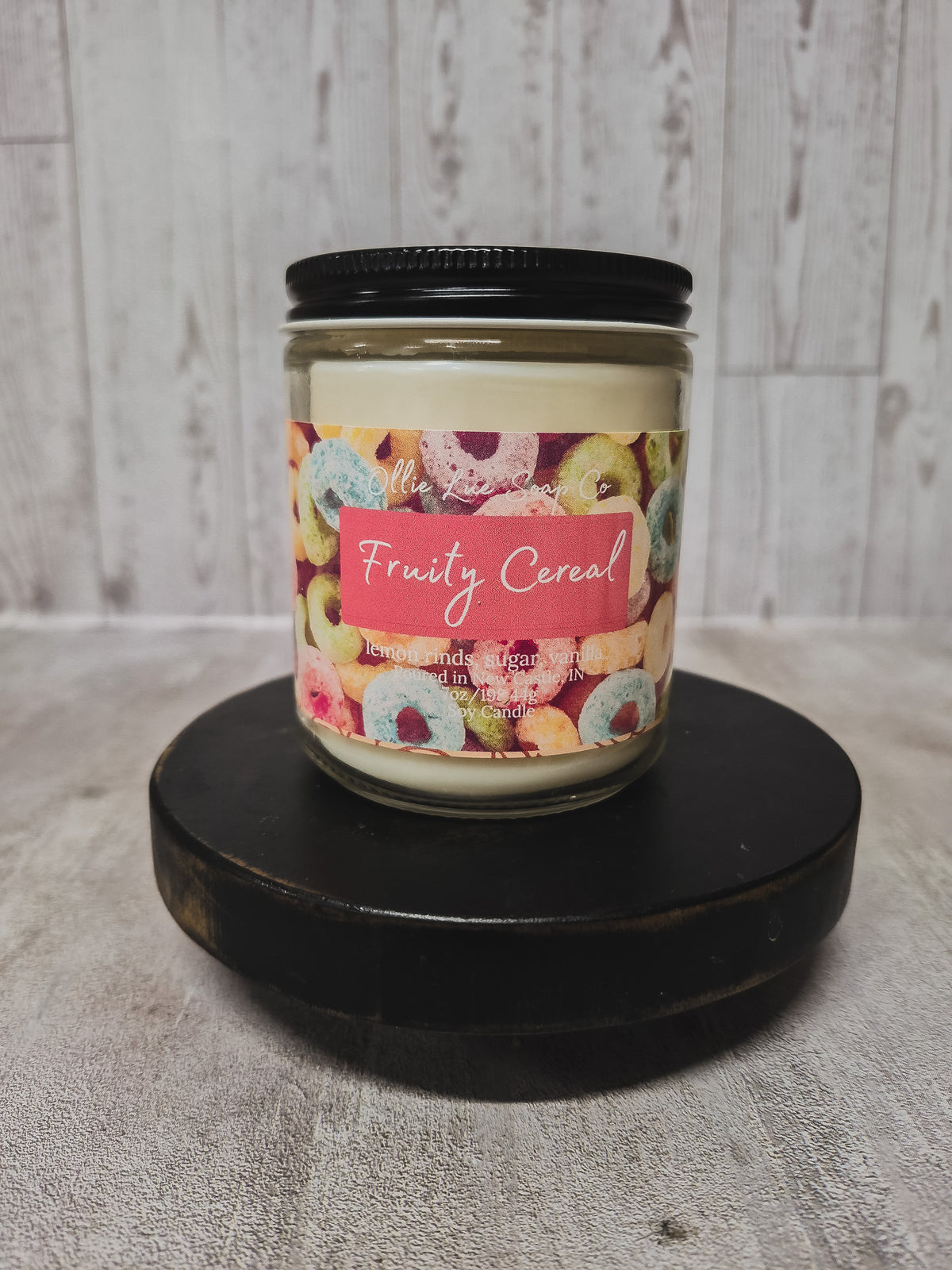 A glass jar candle with a black lid and a colorful donut-patterned label reading 'Fruity Cereal,' displayed on a round black pedestal against a light wood plank background.