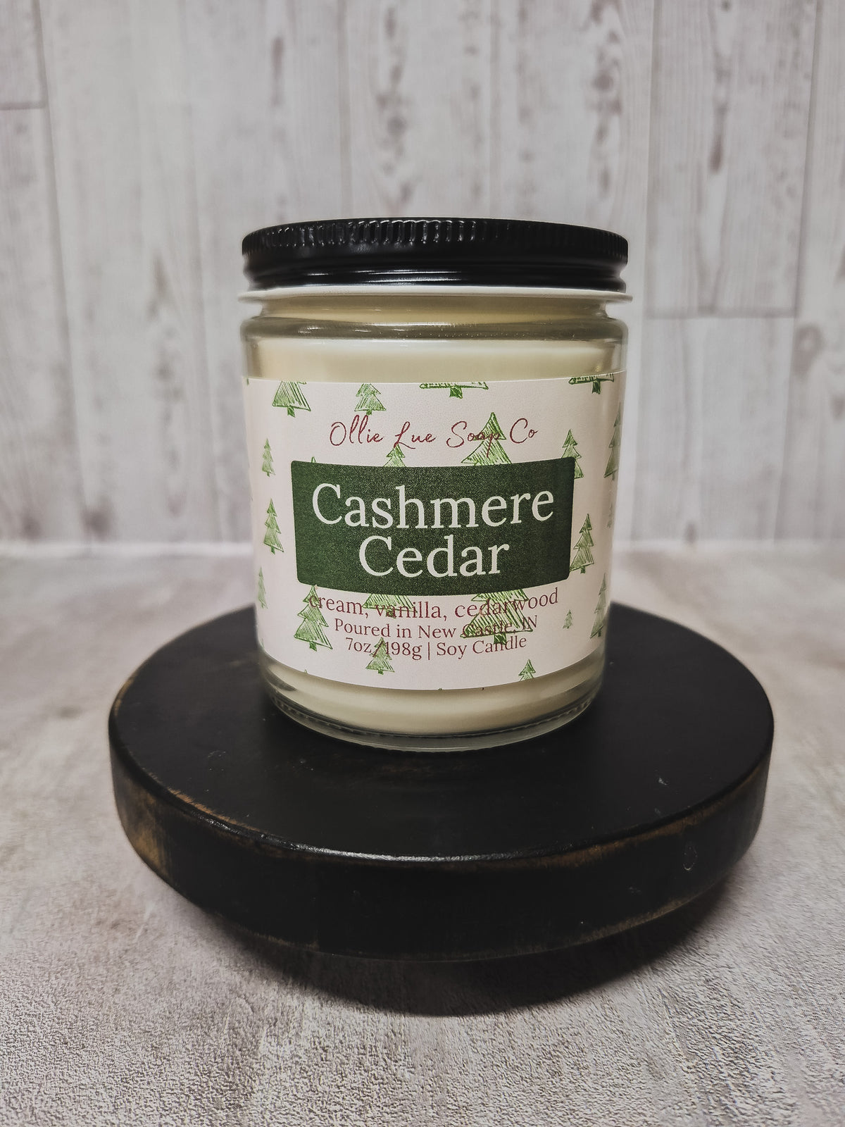 A jar candle labeled 'Cashmere Cedar' by Ollie Lue Soap Co., shown on a round black wooden riser against a light gray textured backdrop and whitewashed wood wall.