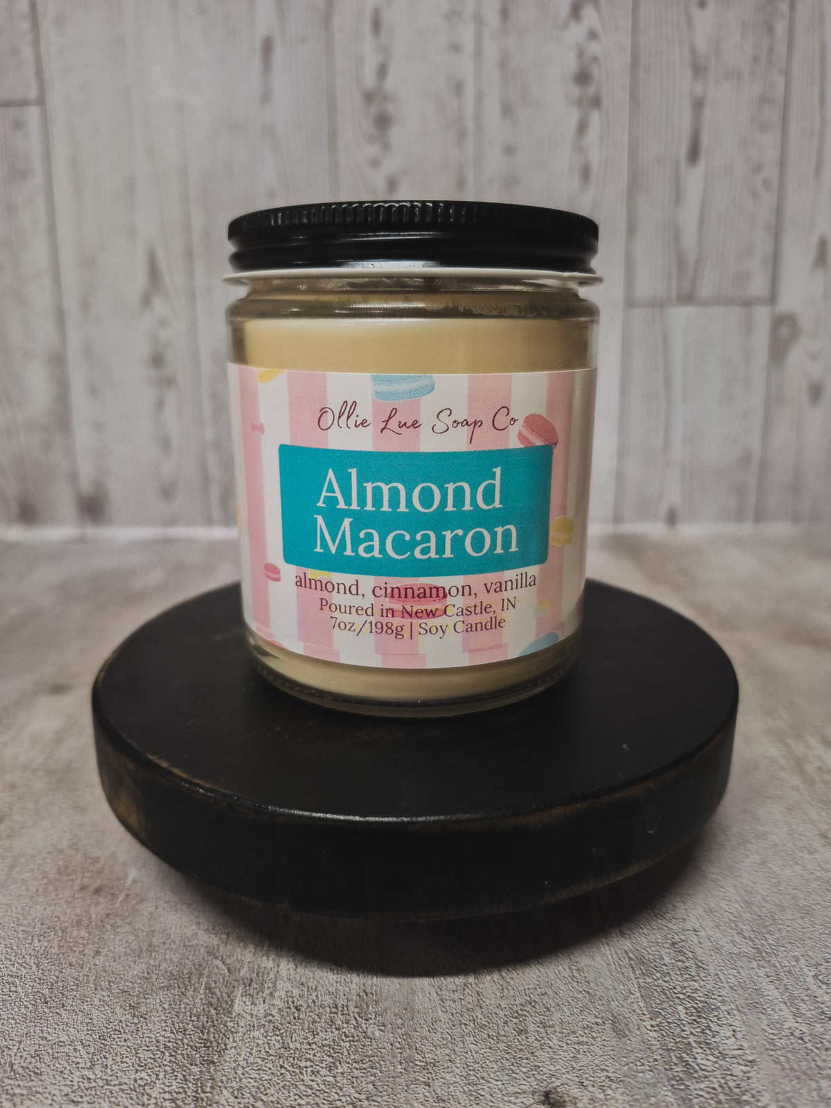 A jar of Ollie Lue Soap Co. Almond Macaron scented soy candle, displayed on a black circular pedestal against a light gray wood-paneled background.