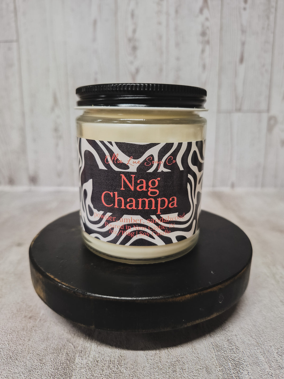 A jar candle labeled 'Nag Champa' by Ollie Lue Soap Co, featuring a black and white zebra-patterned label with red text, set on a round black wooden pedestal against a light gray wood plank background.