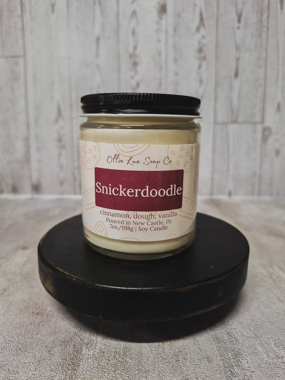 A jar candle labeled 'Snickerdoodle' by Ollie Lue Soap Co, placed on a round black wooden platform against a light gray wood plank background.