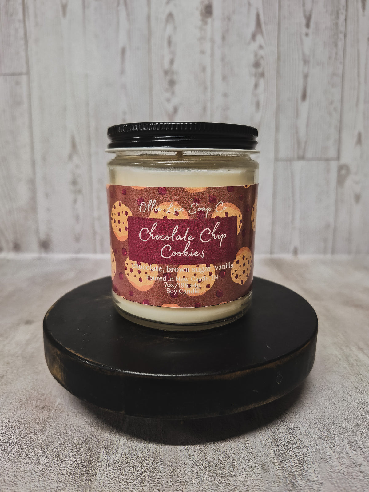 A jar candle labeled 'Ollie Lue Soap Co Chocolate Chip Cookies' sits on a round black wooden platform against a light gray wood-plank background.