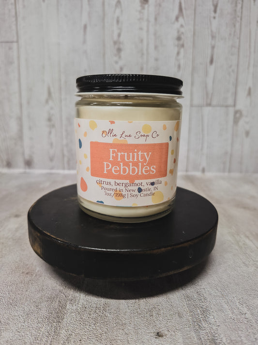 a candle by Ollie lue Soap co in a clear jar labeled Fruity Pebbles on a black stool in a gray background